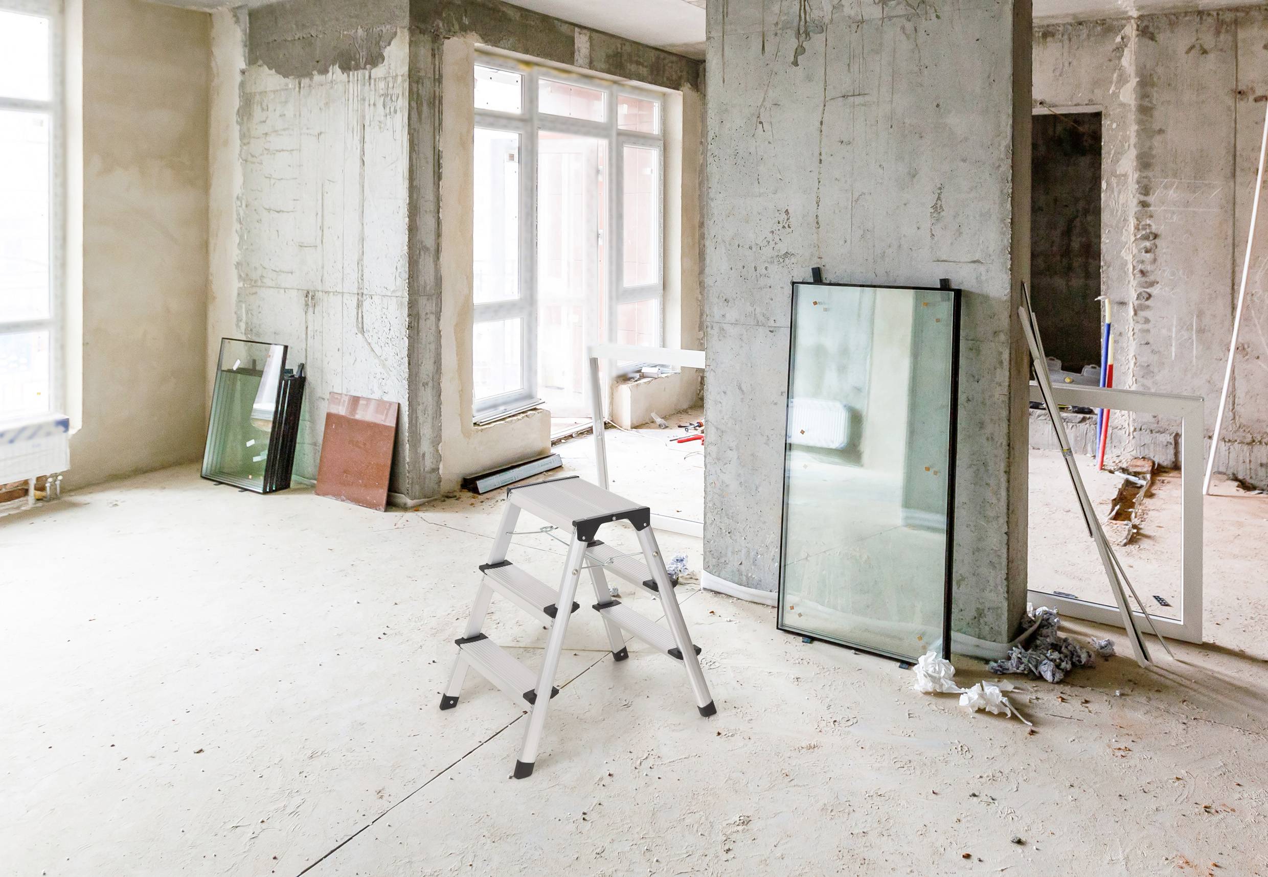 Ein leerer Raum in einem Rohbau mit einer Aluminiumleiter und mehreren Glasplatten an der Wand. Tageslicht strömt durch Fenster.