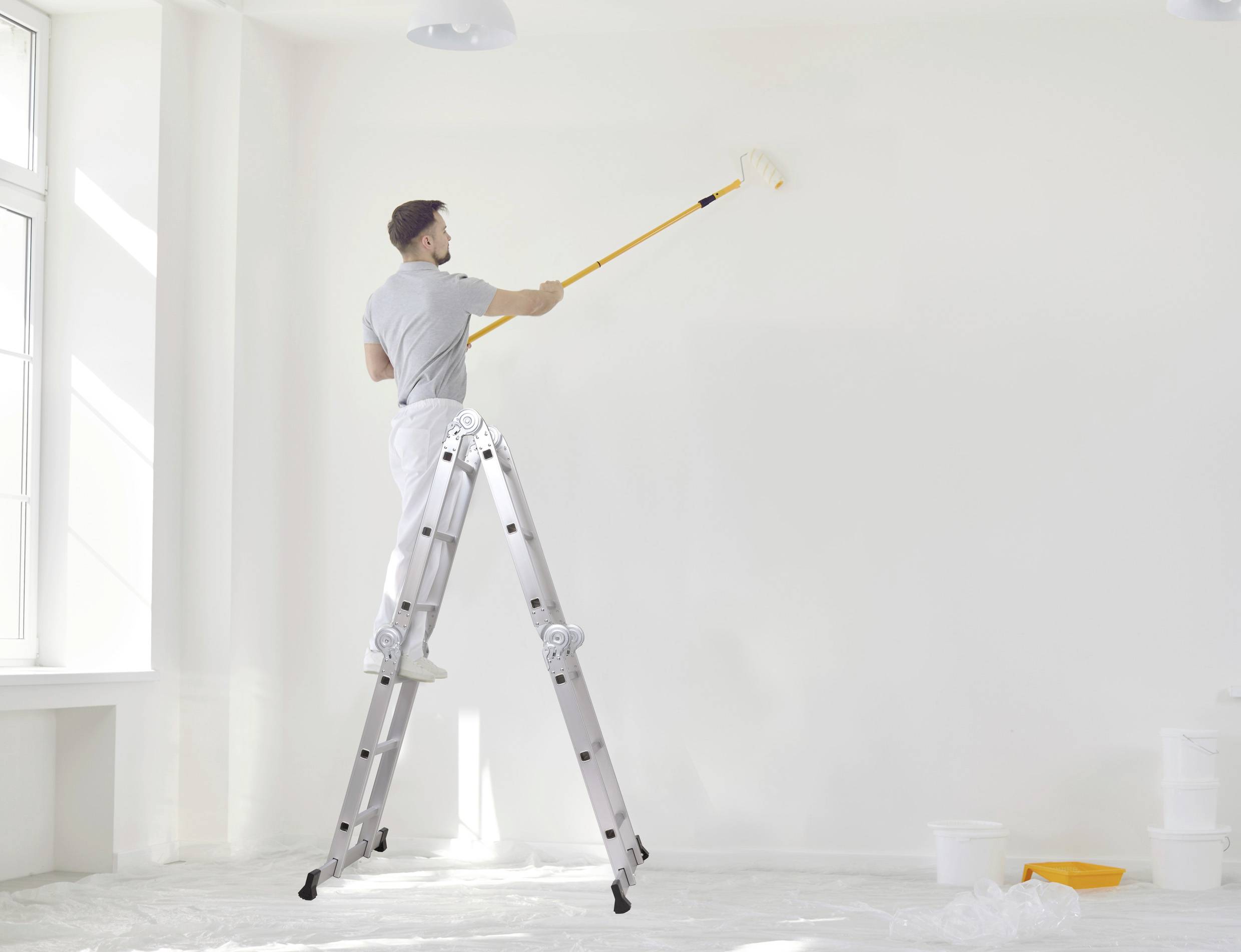 Ein Mann steht auf einer Leiter und streicht eine weiße Wand mit einer Farbrolle in einem hellen, gut beleuchteten Raum.