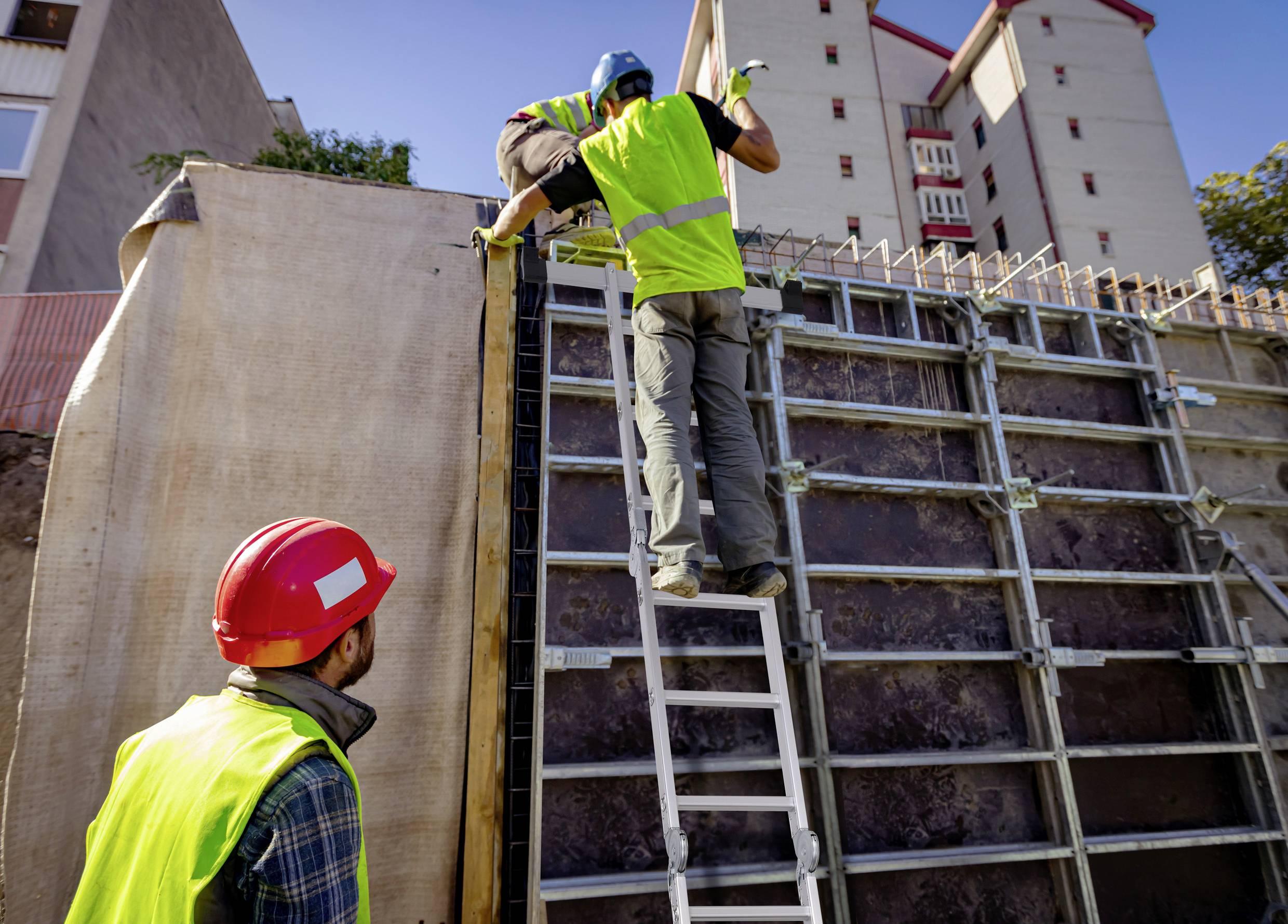 Arbeiter in Sicherheitskleidung errichten eine Mauer mit Hilfe einer Leiter auf einer Baustelle vor einem mehrstöckigen Gebäude.