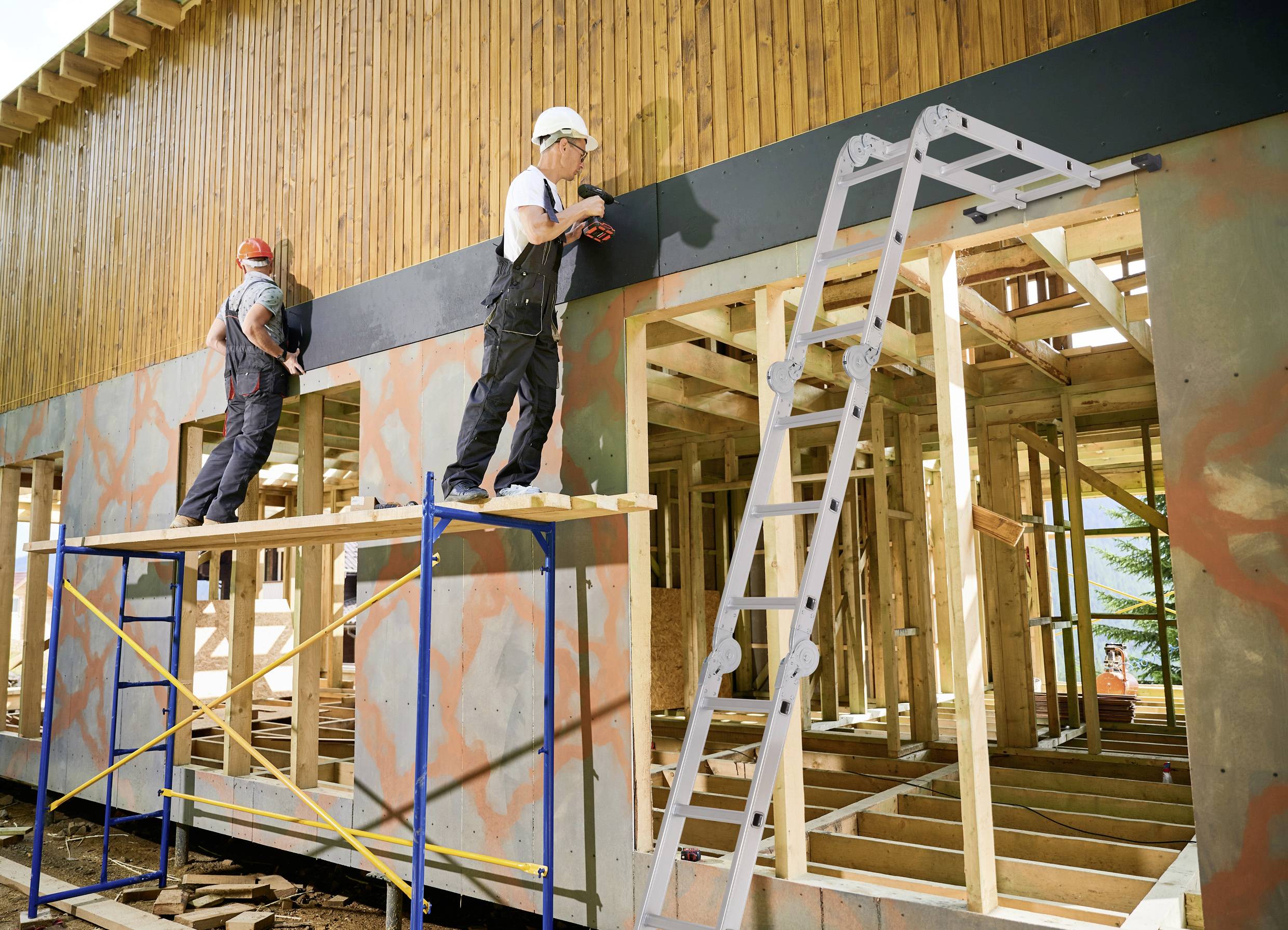 Zwei Bauarbeiter renovieren die Fassade eines Hauses. Einer steht auf einem Gerüst, der andere auf einer Leiter. Das Gebäude hat eine Holzwand.