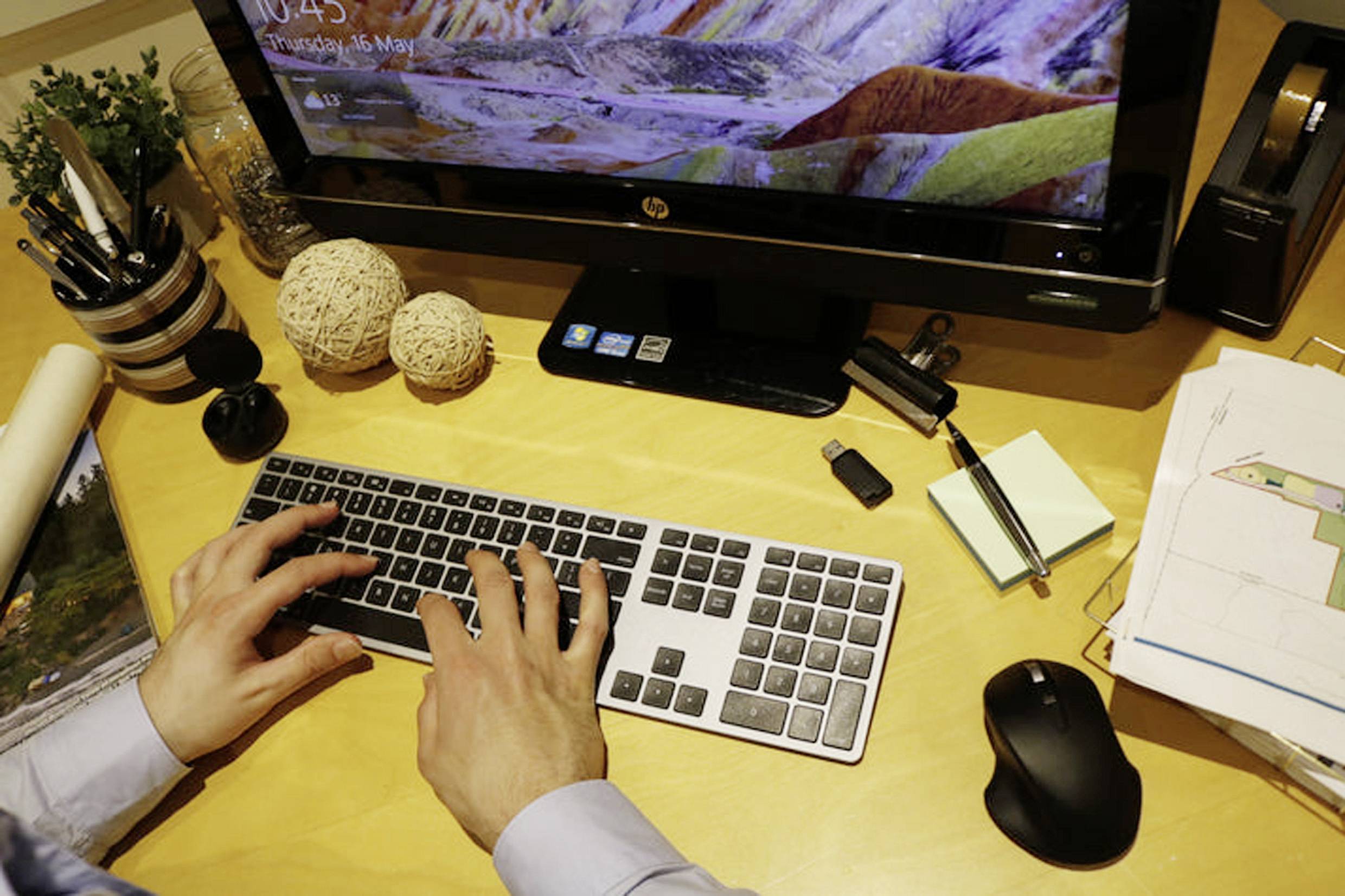 Person tippt auf einer Tastatur an einem Schreibtisch. Auf dem Bildschirm ist ein buntes Landschaftsbild zu sehen. Büroartikel liegen verstreut.