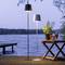 Ein Tisch mit Getränken steht auf einer Terrasse am See, flankiert von zwei modernen Stehlampen. Im Hintergrund ist das Wasser zu sehen.