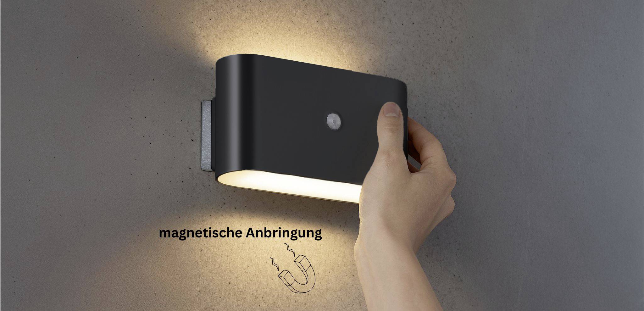 Eine Hand justiert eine schwarze Wandleuchte und zeigt ihre magnetische Befestigung. Die Leuchte verbreitet ein warmes Glühen an der grauen Wand. Der Text 'Magnetische Anbringung' mit einem Magneticon ist sichtbar.