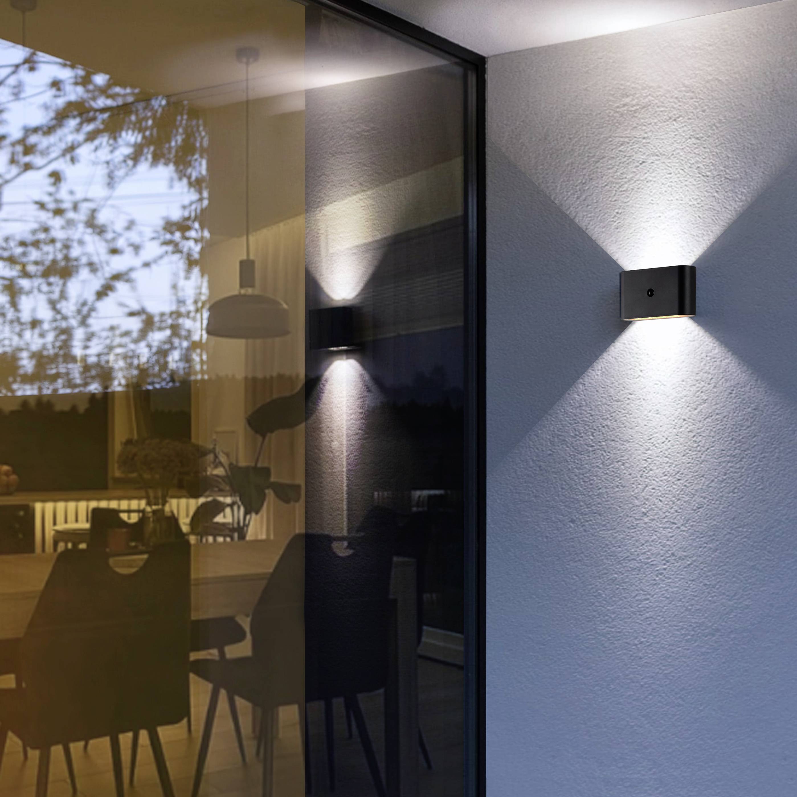 Eine moderne Wandleuchte beleuchtet die Außenseite eines Hauses. Durch das große Glasfenster ist ein Esszimmer mit Stühlen und einem Tisch zu sehen.