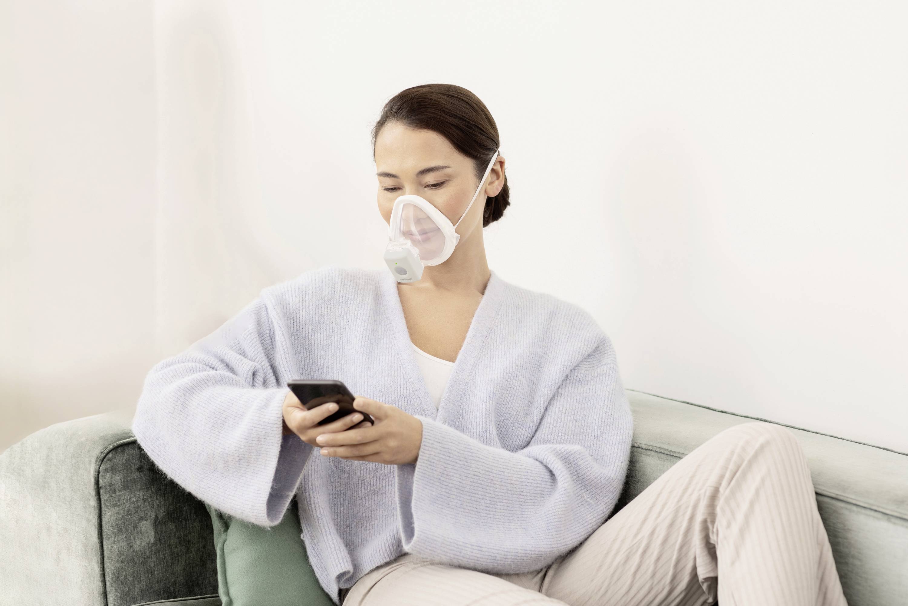 Eine Person sitzt auf einem Sofa, trägt eine Atemschutzmaske und benutzt ein Smartphone.