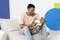 Mann mit rosa T-Shirt und Jeans sitzt auf Sofa und betrachtet ein Gerät in seiner Hand. Farbige Formen im Hintergrund.