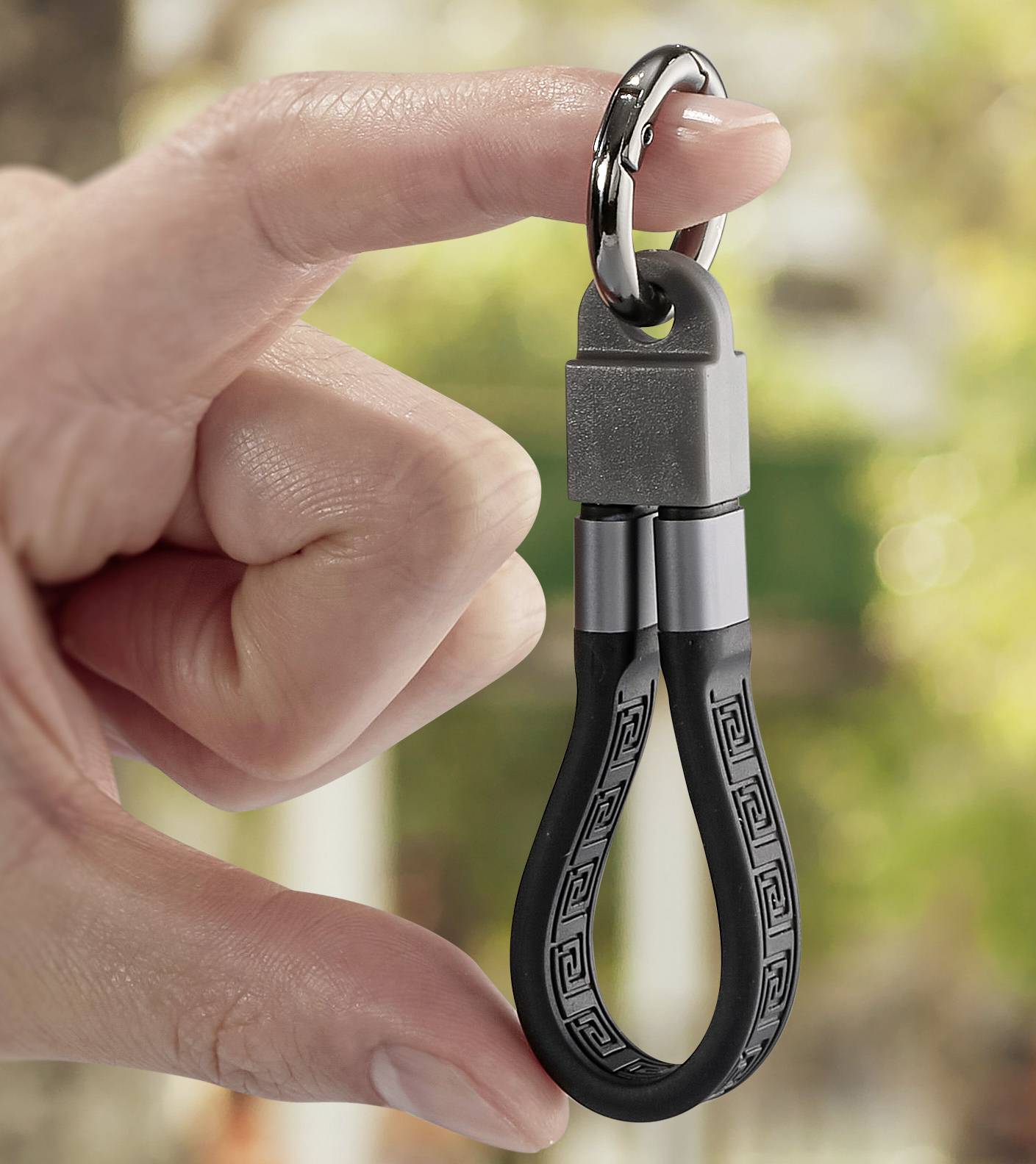 Finger hält einen modernen Schlüsselanhänger mit schwarzem Kabel und Metallverschluss vor unscharfem, grünem Hintergrund.