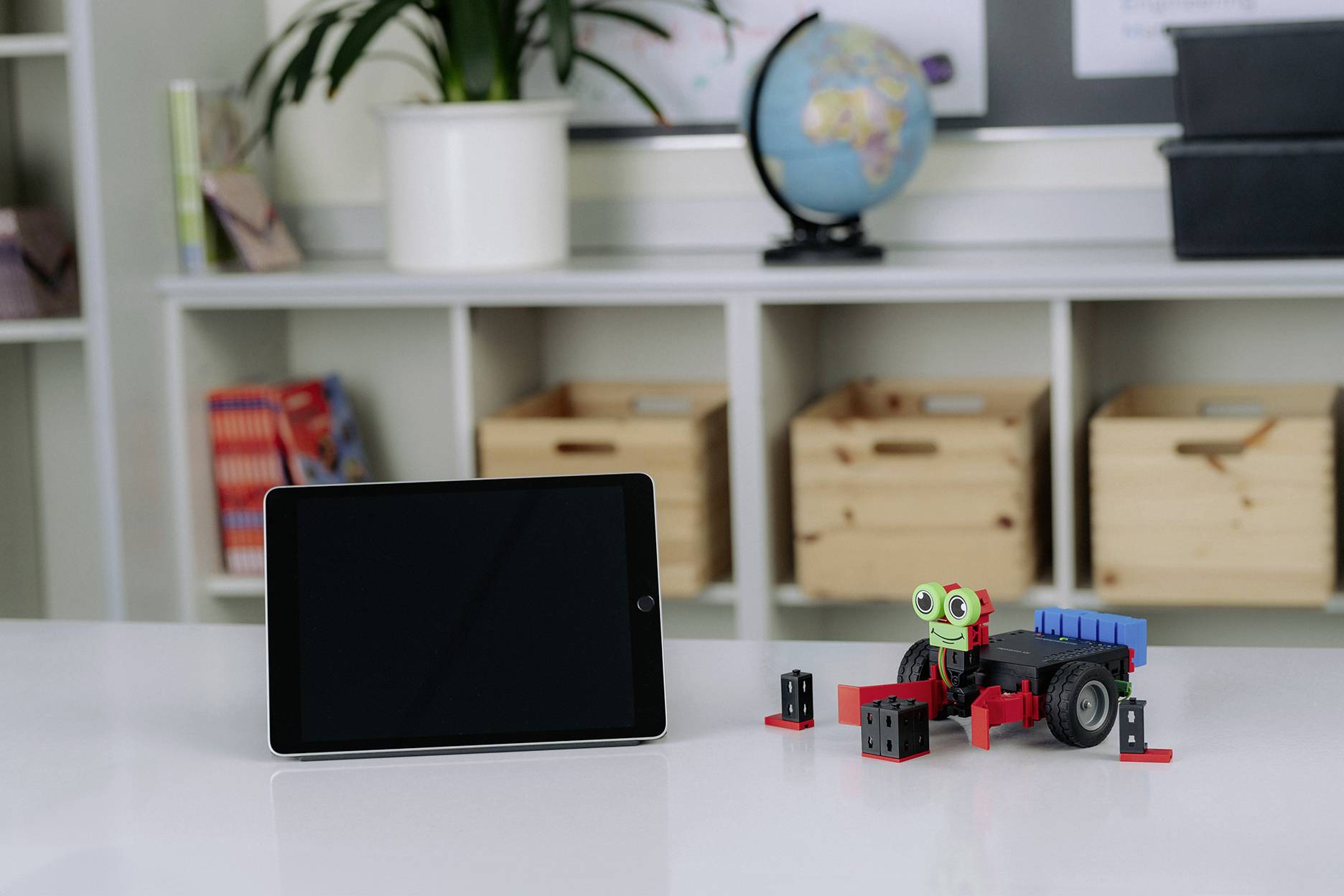 Tablet neben einem kleinen Roboterfahrzeug aus bunten Bausteinen auf einem Tisch, mit Büchern und einem Globus im Hintergrund.