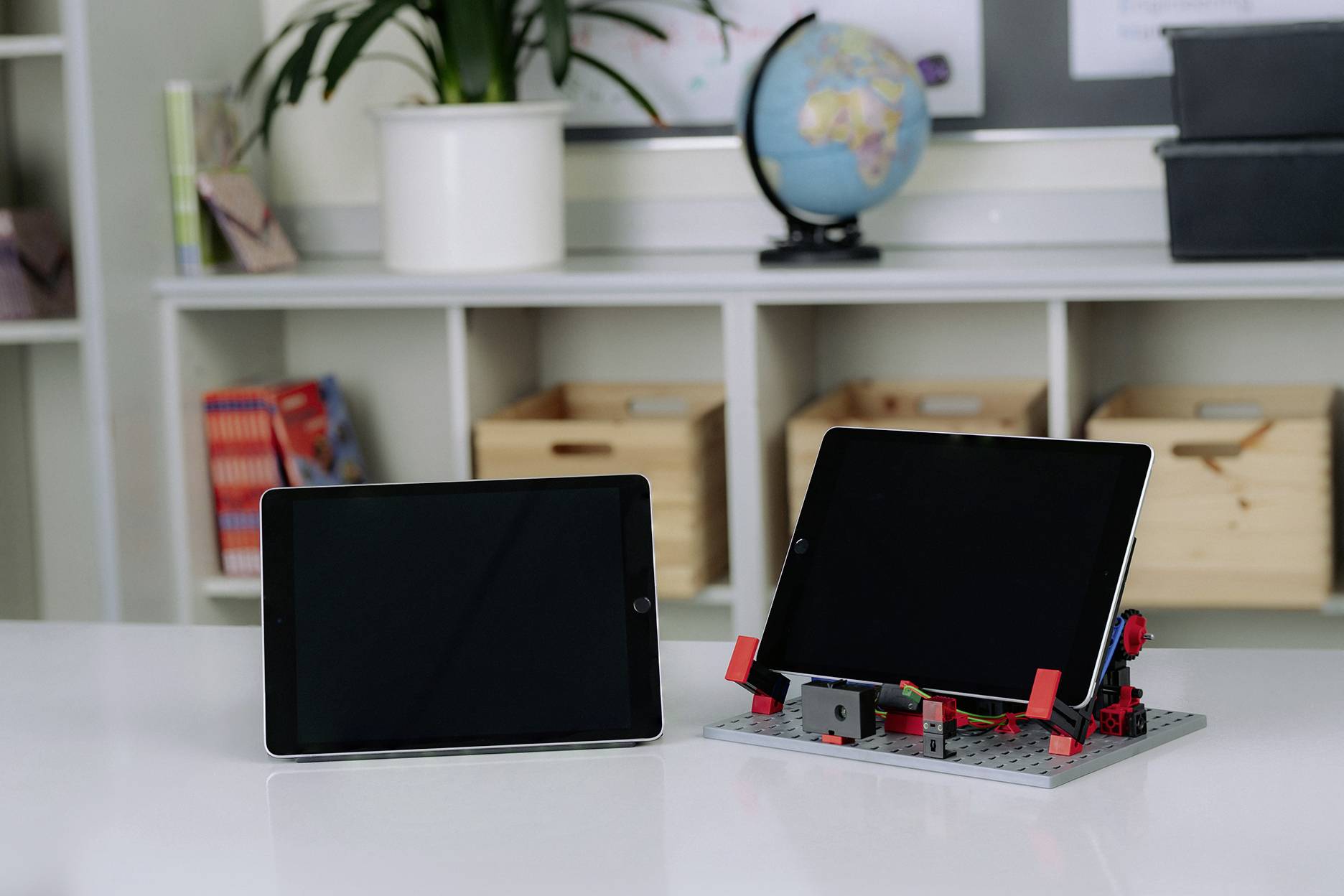 Zwei Tablets auf einem Tisch, eines liegt flach, das andere ist in einem Lego-Ständer aufrecht positioniert. Im Hintergrund ein Globus.