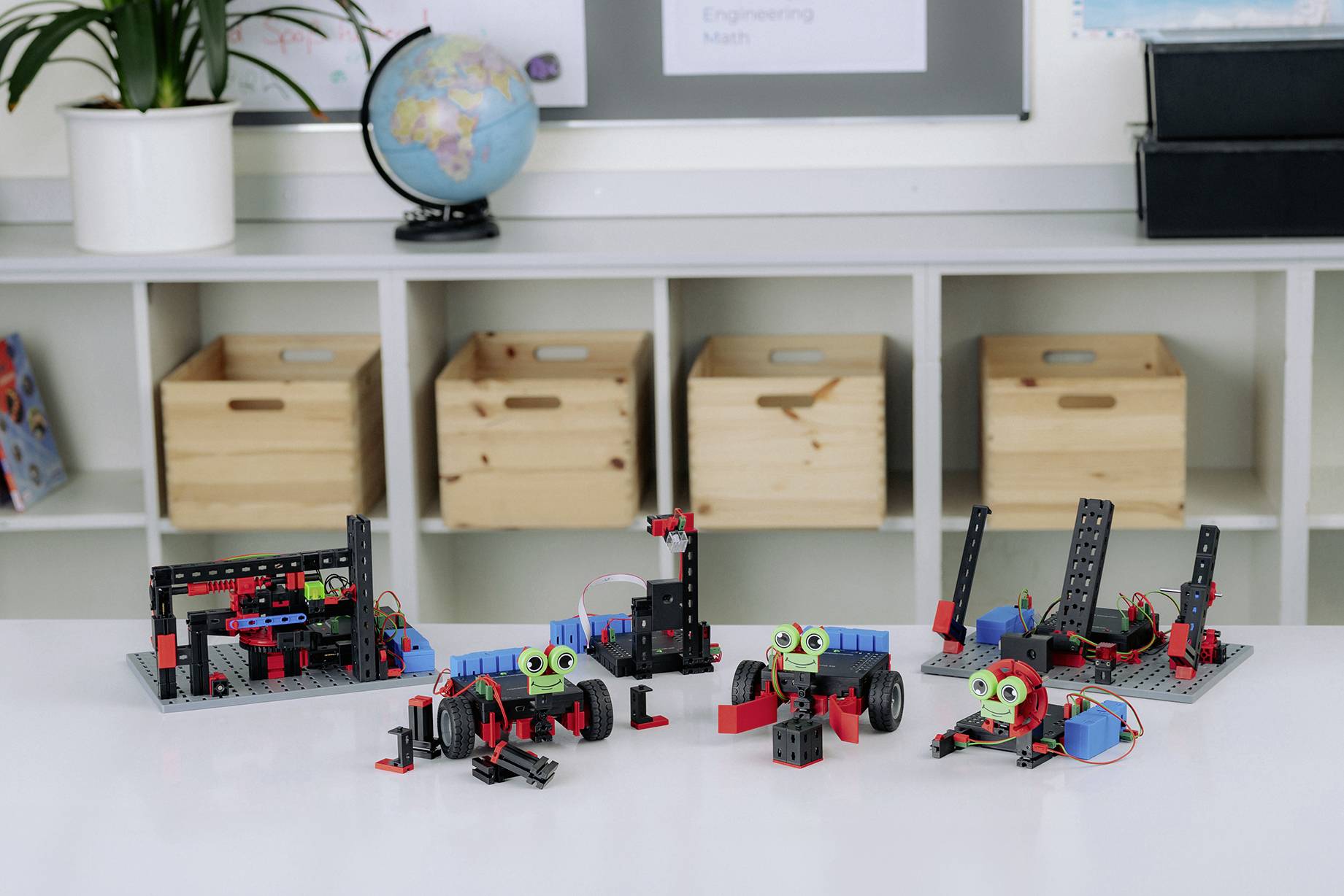 Auf einem Klassenzimmer-Tisch stehen mehrere kleine Roboter aus Bauklötzen, Sensoren und Motoren. Im Hintergrund eine Weltkugel und Holzboxen.