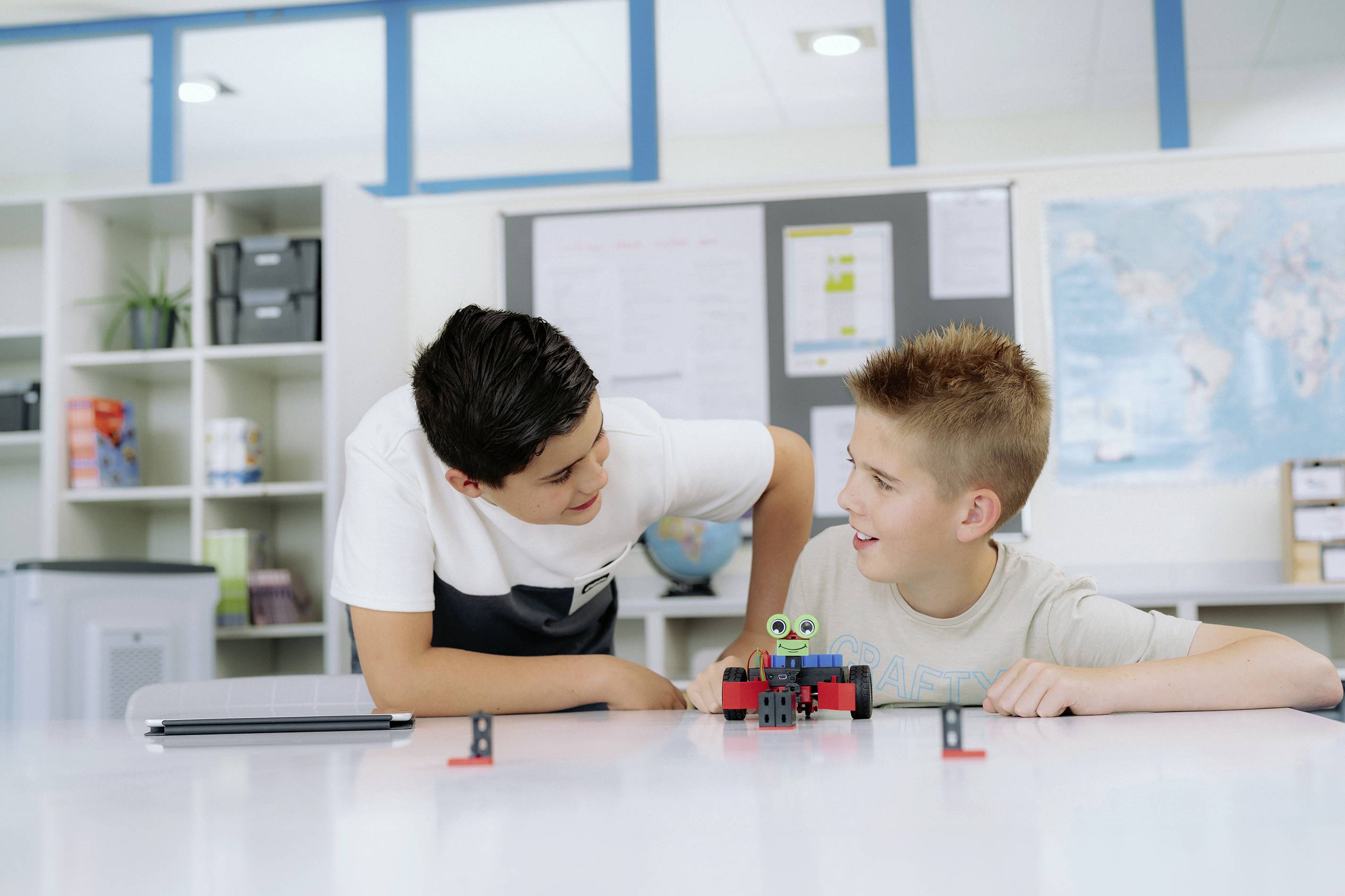 Zwei Jungen bauen gemeinsam ein kleines Roboterfahrzeug im Klassenzimmer. Einer zeigt auf etwas, während der andere ihn ansieht und lacht.