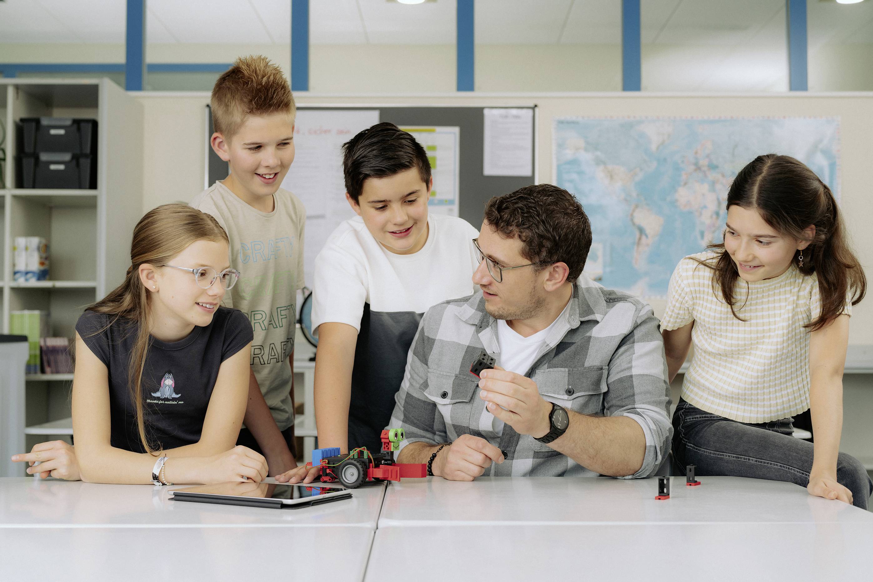 Eine Gruppe von Kindern sitzt mit einem Erwachsenen an einem Tisch. Der Erwachsene zeigt den Kindern ein kleines Modellfahrzeug. Im Hintergrund ein Büroschrank und eine Weltkarte.