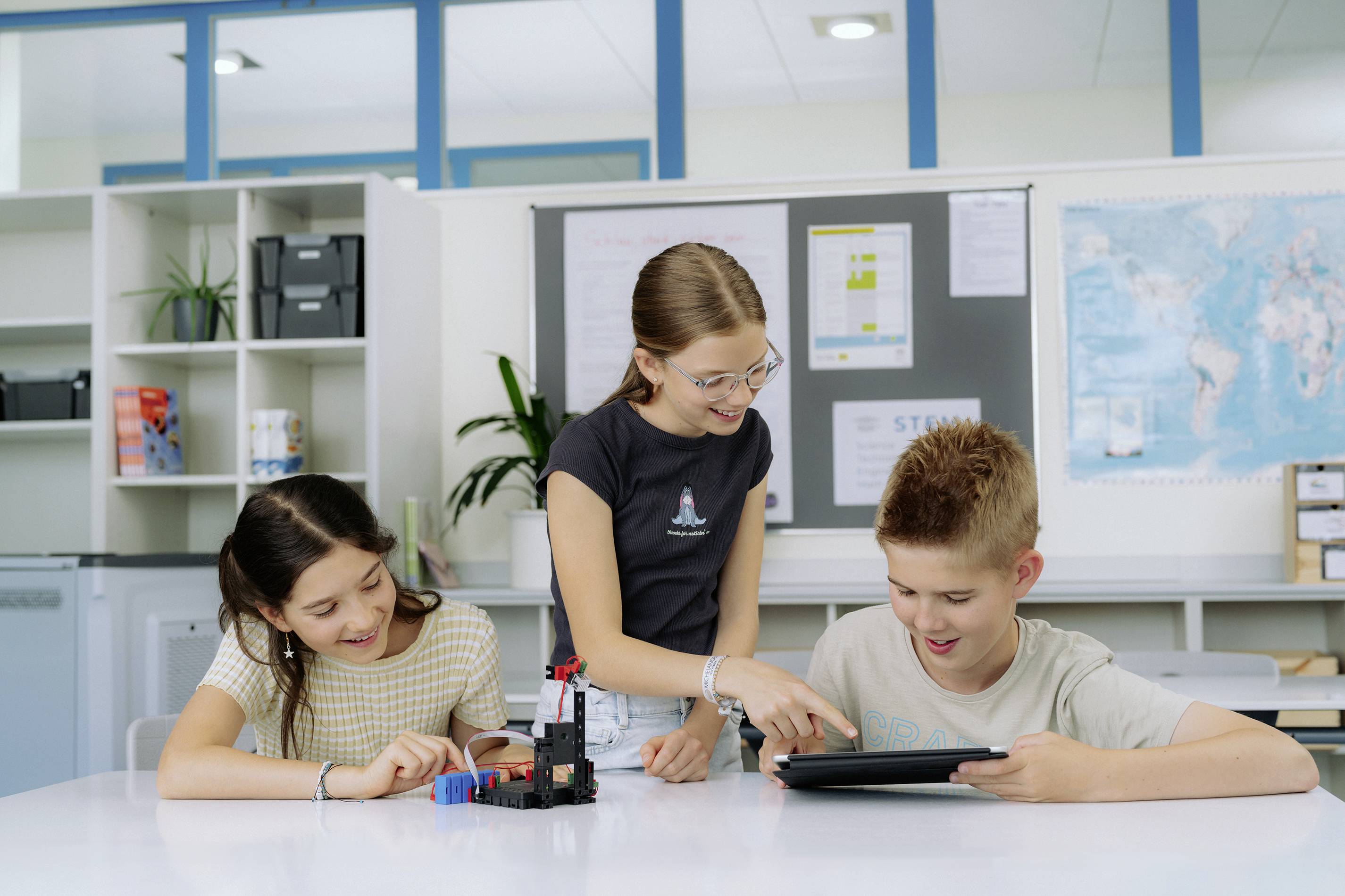 Drei Kinder arbeiten gemeinsam an einem Tisch in einem modernen Klassenraum. Sie verwenden ein Tablet und haben einen kleinen Roboter vor sich.