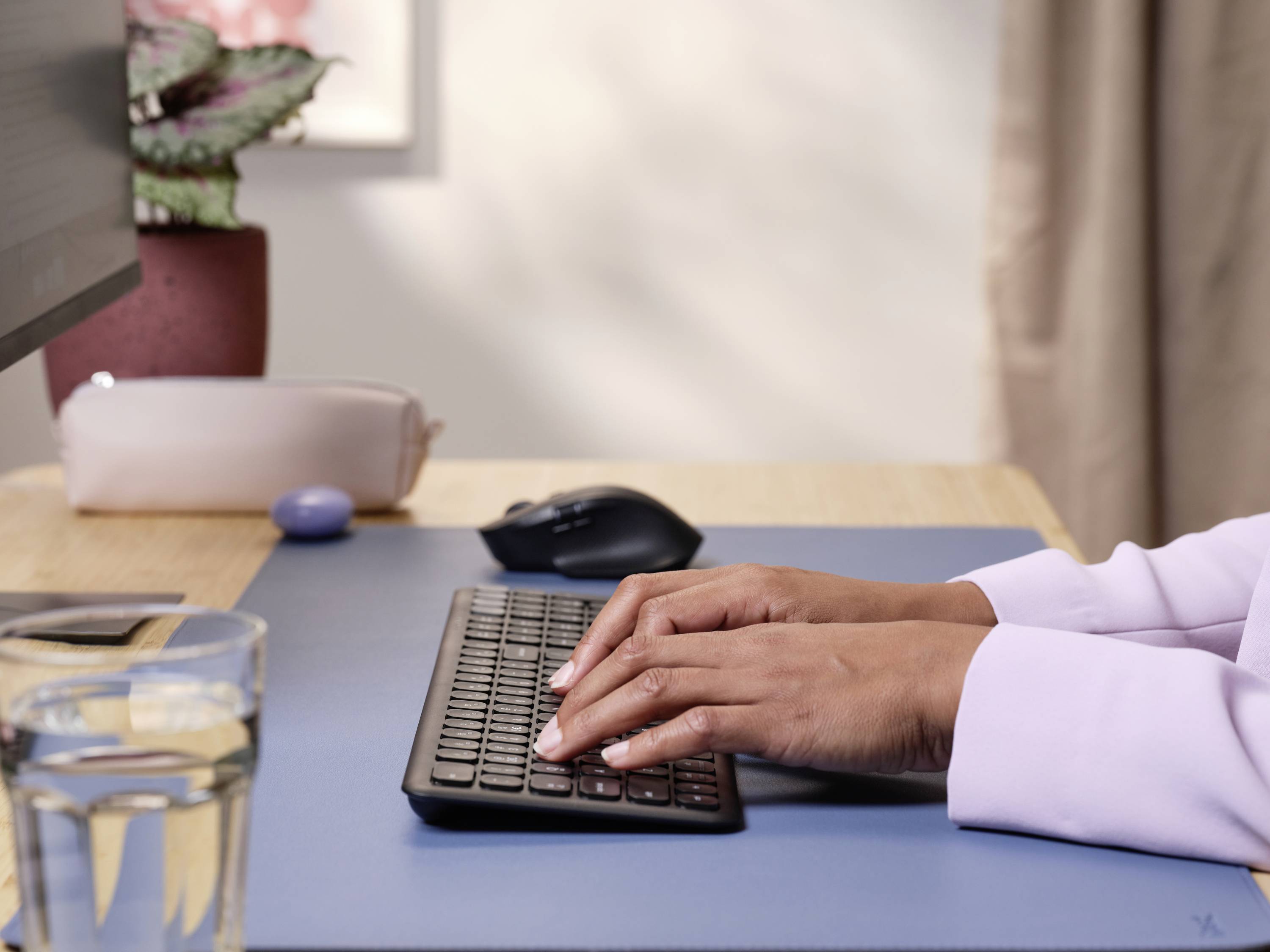 Person tippt auf einer Tastatur an einem Schreibtisch, auf dem ein Glas Wasser, Computermaus und eine Pflanze stehen.