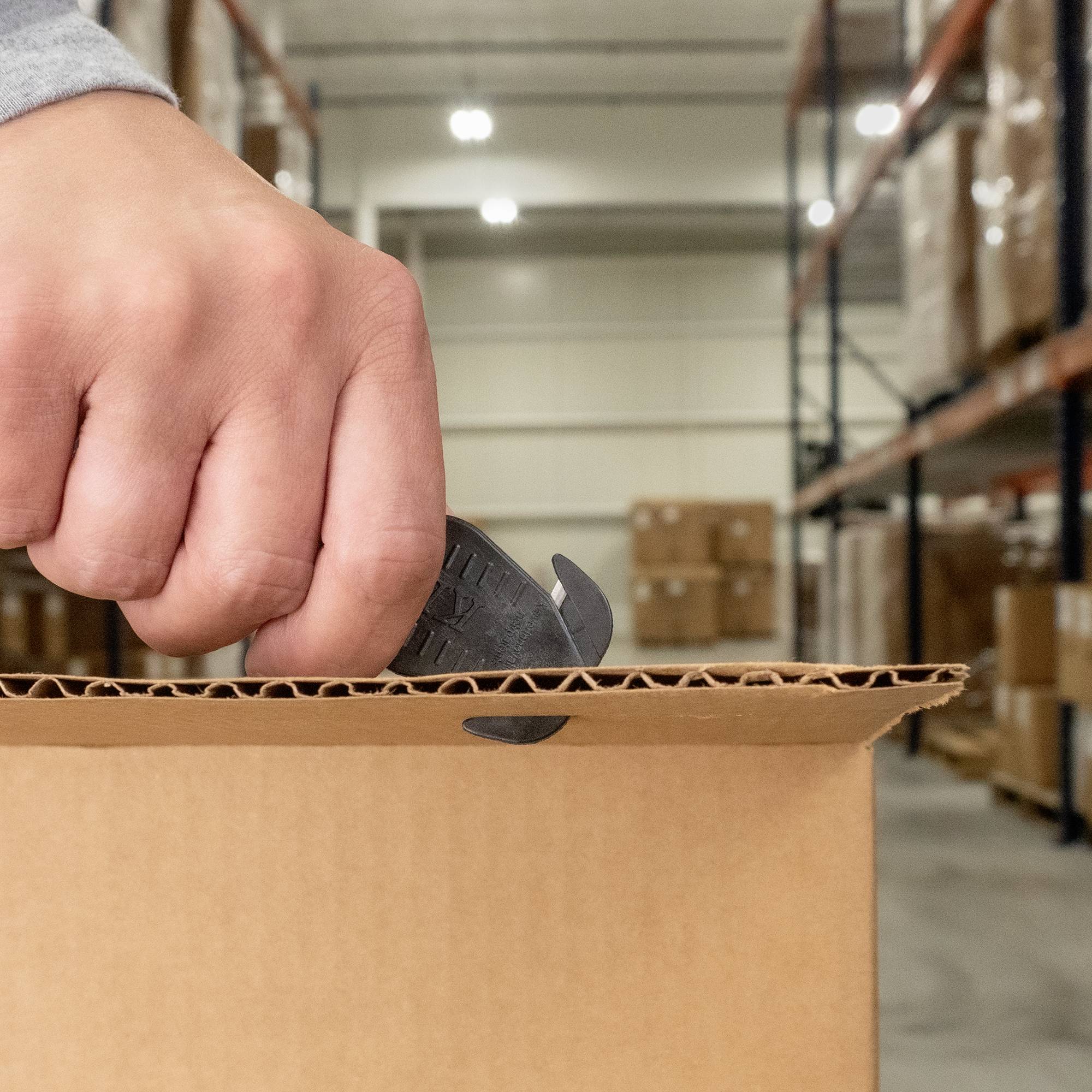 Eine Person öffnet in einem Lager mit einem Cutter-Messer einen Karton. Im Hintergrund sind Regale mit weiteren Kartons zu sehen.