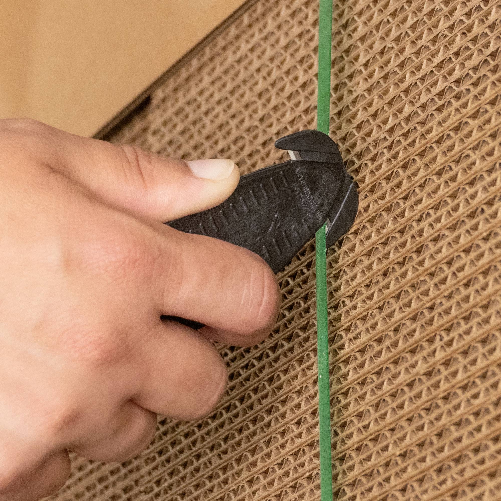 'Nahaufnahme einer Hand, die mit einem Cutter-Messer eine grüne Plastikschnur auf einer braunen Wellpappe durchschneidet.'