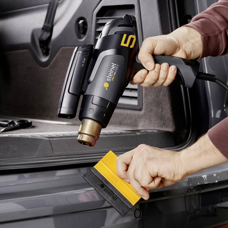 Eine Person bedient ein Heißluftgerät mit digitalem Display an einem Fahrzeug. Sie hält ein gelbes Werkzeug in der anderen Hand.