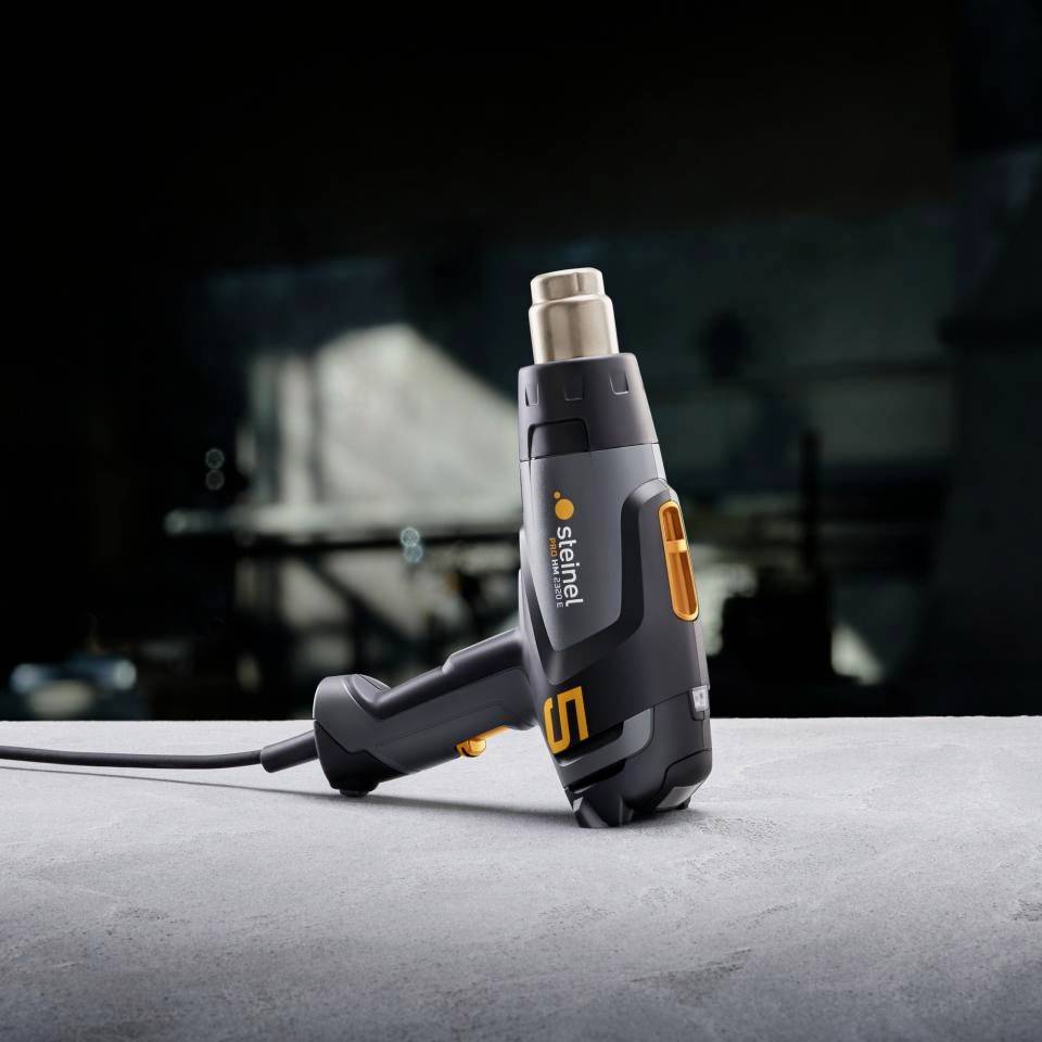 Ein elektrischer Heißluftföhn liegt auf einem Tisch in einer Werkstatt. Im Hintergrund sind unscharfe Werkzeuge und Werkbank erkennbar.