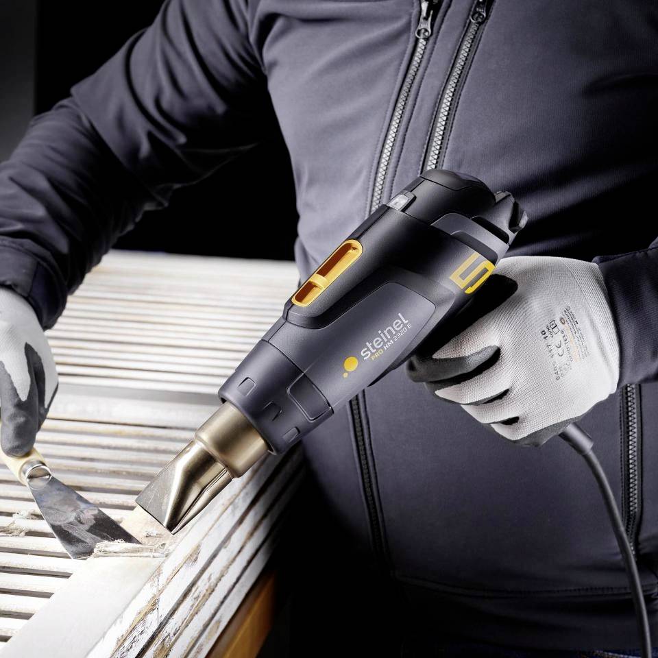 Person verwendet eine Heizpistole, um Farbe von einer Holzoberfläche zu entfernen. Sie trägt Arbeitshandschuhe und eine graue Jacke.