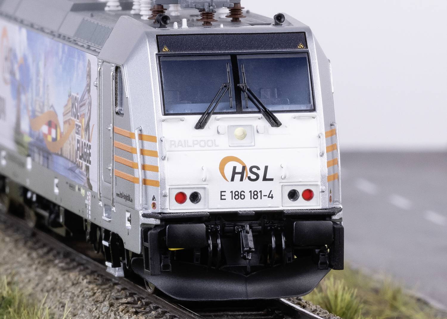 Elektrolokomotive in silberner Farbe mit HSL-Logo und der Nummer 'E 186 181-4', fährt auf Gleisen, gezeigt von vorne.