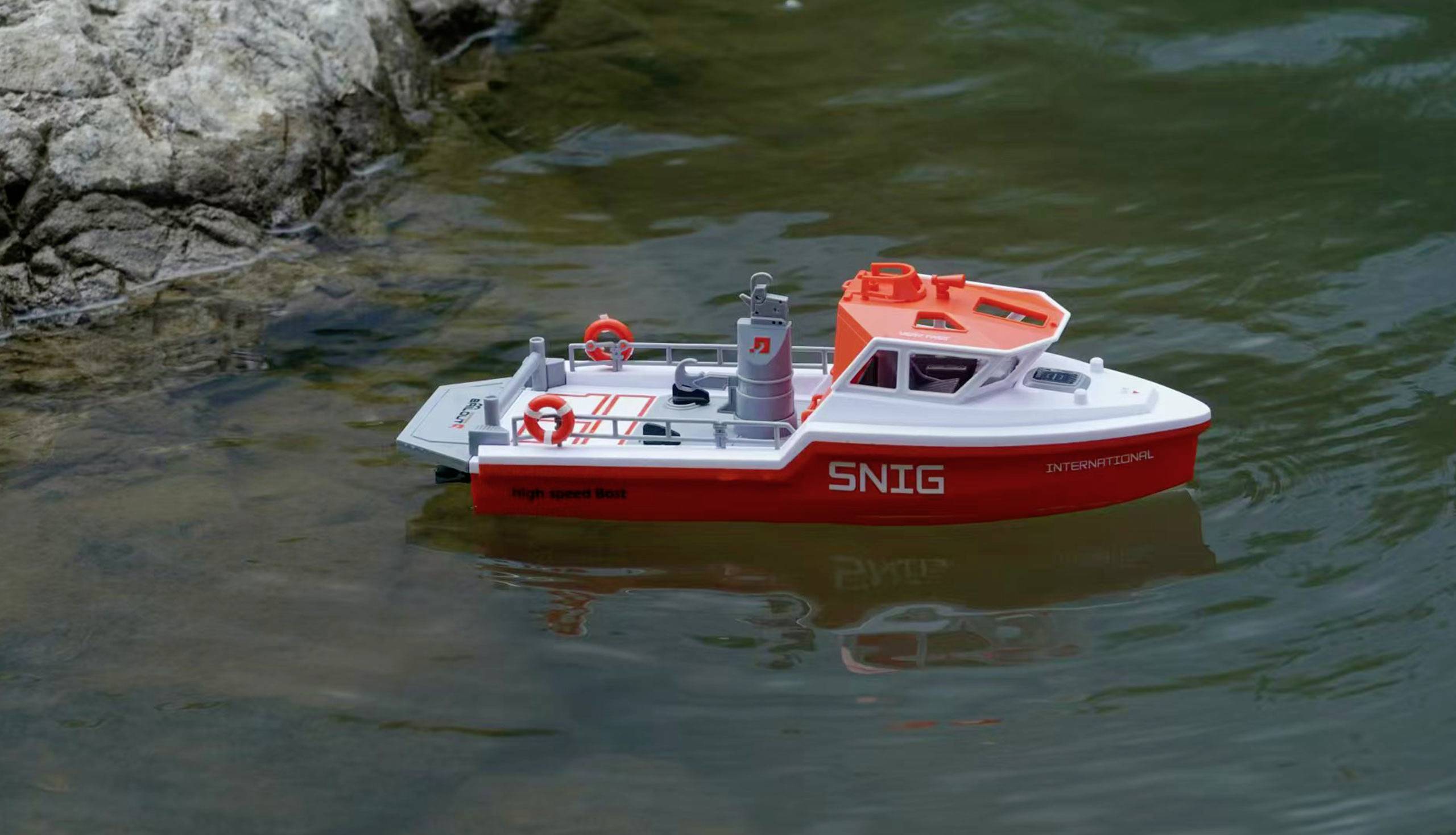 Ein kleines Modellboot mit der Aufschrift 'SNIG' schwimmt im Wasser nahe einem Felsen. Das Boot hat rote und weiße Farben.
