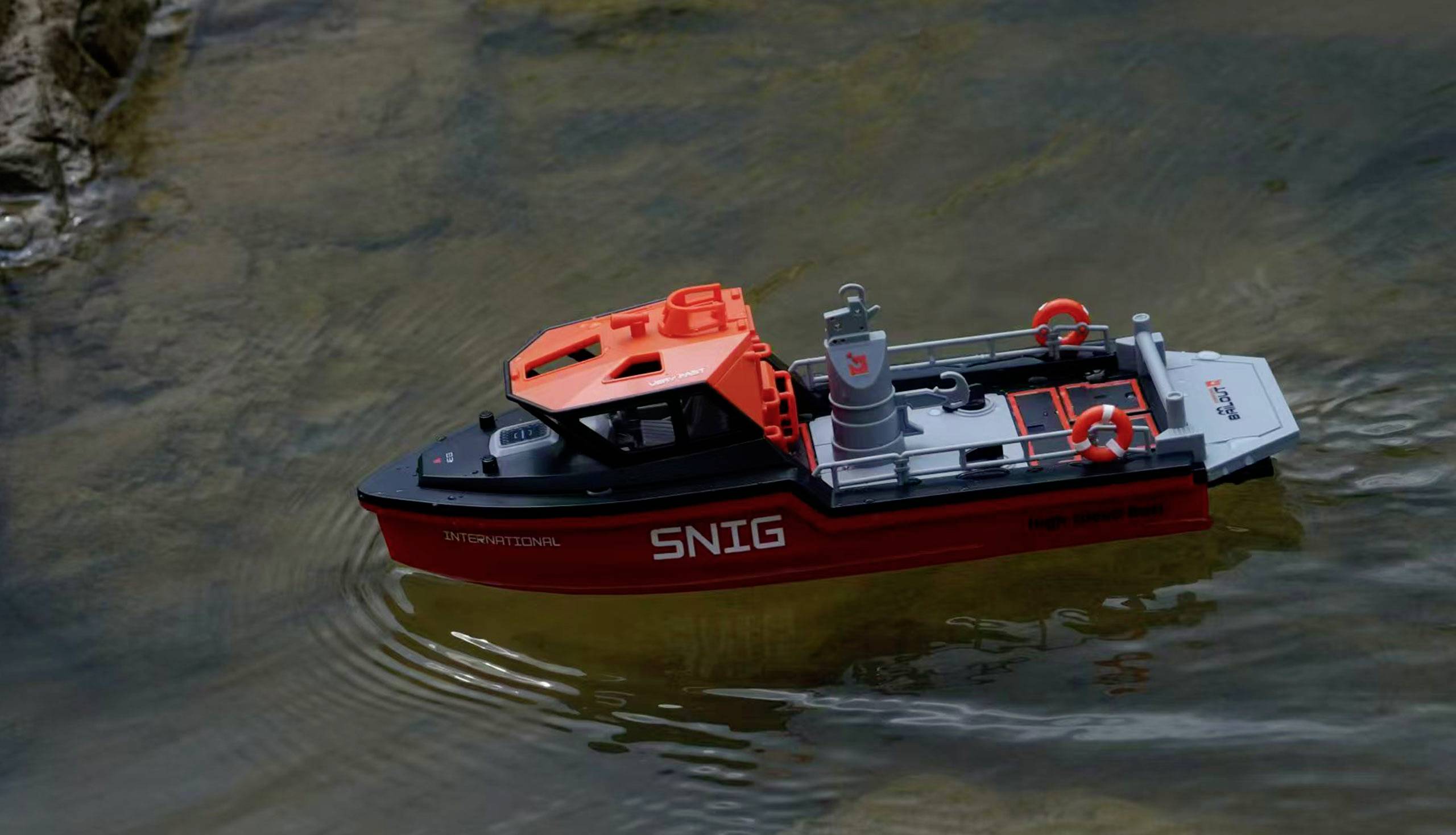 Ein kleines Modellboot mit der Aufschrift 'SNIG' fährt auf ruhigem Wasser nahe einem steinigen Ufer.