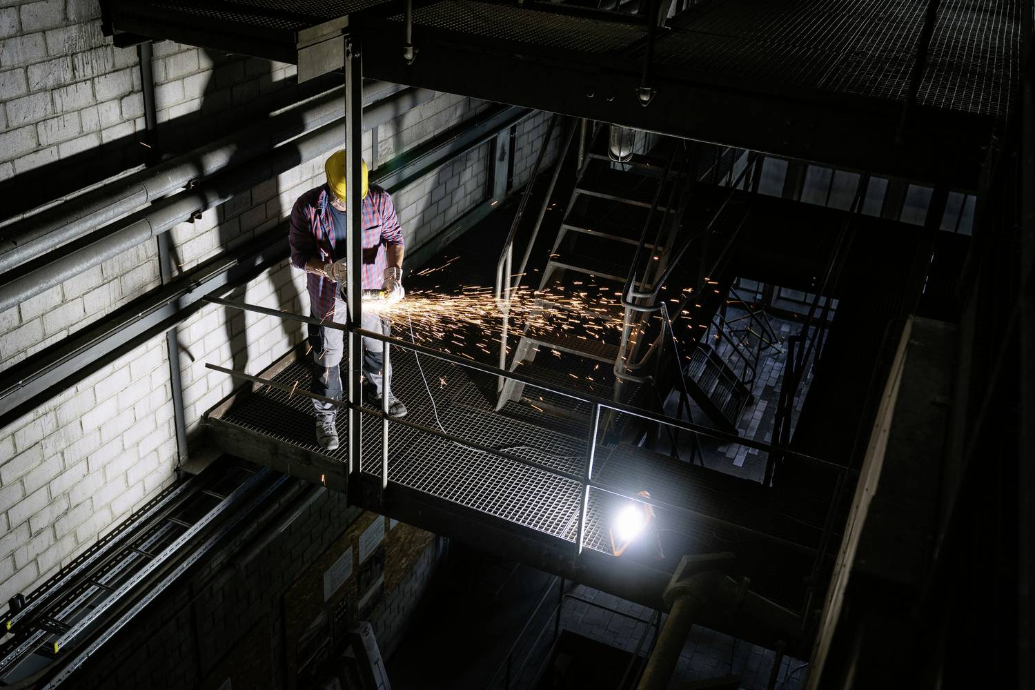 Ein Arbeiter mit Helm schweißt in einer Fabrik, Funken fliegen. Beleuchteter Industriebereich mit Metallkonstruktionen im Hintergrund.