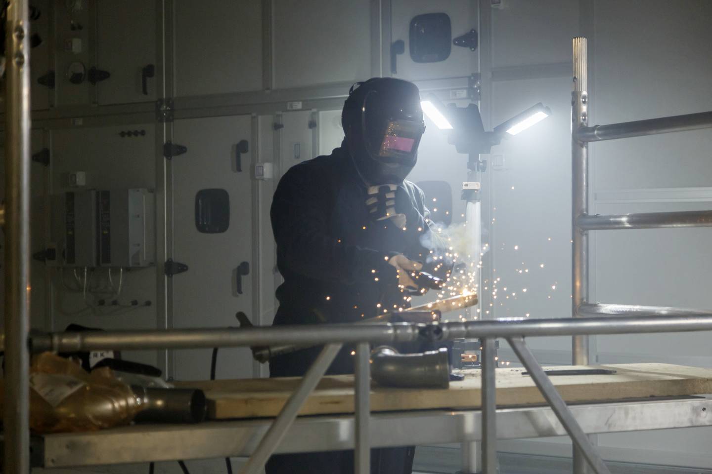 Ein Schweißer, der einen Schutzanzug und Helm trägt, arbeitet an einem Metallstück. Funken fliegen in einer Werkstattumgebung.