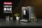 Eine silberne Kaffeemaschine mit 'Crema One Touch'-Anzeige steht auf einer Küchentheke. Daneben sind ein Glas Wasser und ein Kräuterstrauch.