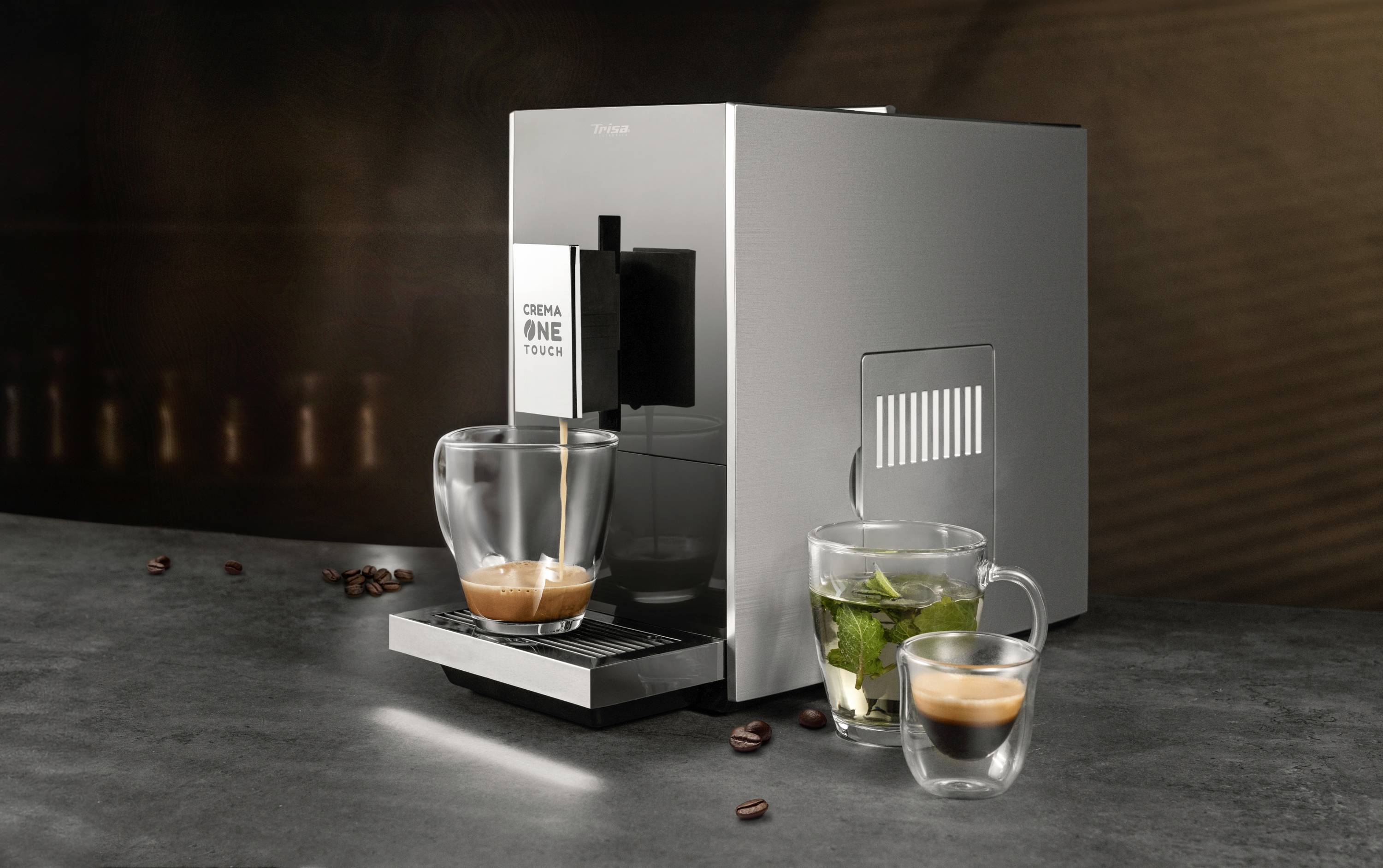 Eine Kaffeemaschine auf einer Küchentheke brüht frischen Espresso. Daneben stehen zwei Gläser, eines mit Tee, eines mit Espresso.