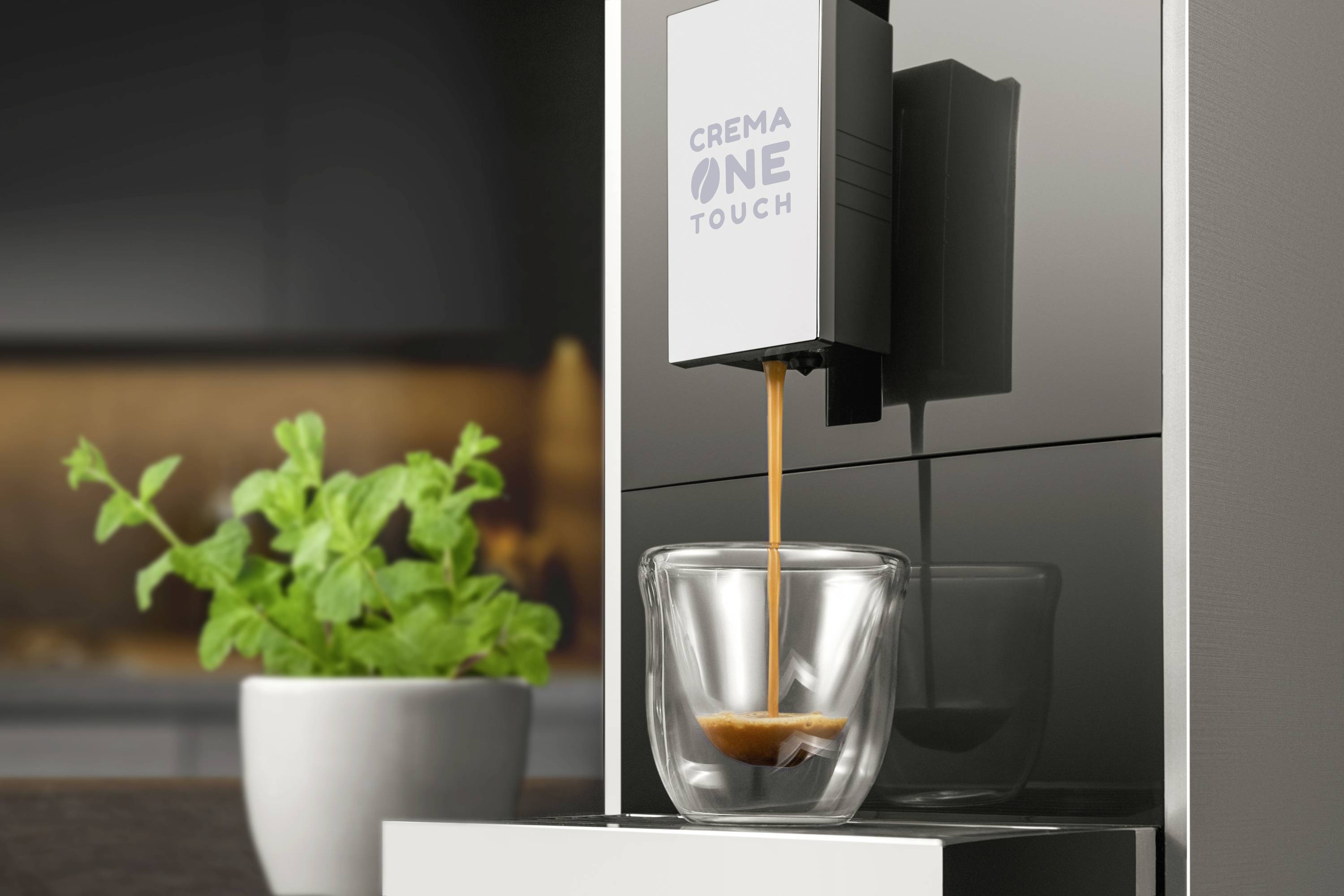 Eine moderne Kaffeemaschine, die Kaffee in eine durchsichtige Tasse brüht. Im Hintergrund steht eine Pflanze in einem weißen Topf.