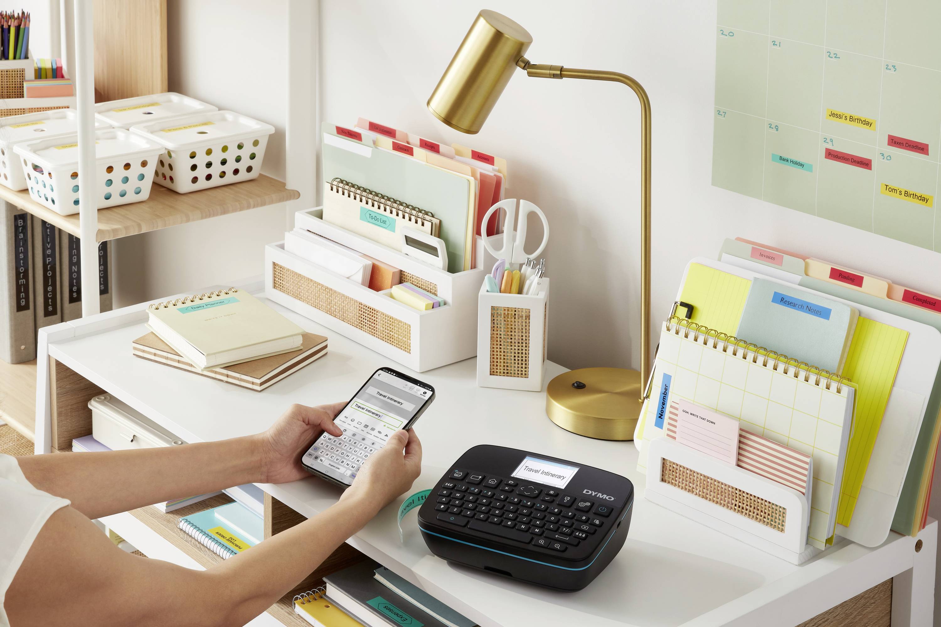 Ein aufgeräumter Schreibtisch mit einem Etikettendrucker, einem Smartphone, Büroaccessoires, einer Schreibtischlampe und Ordnern.