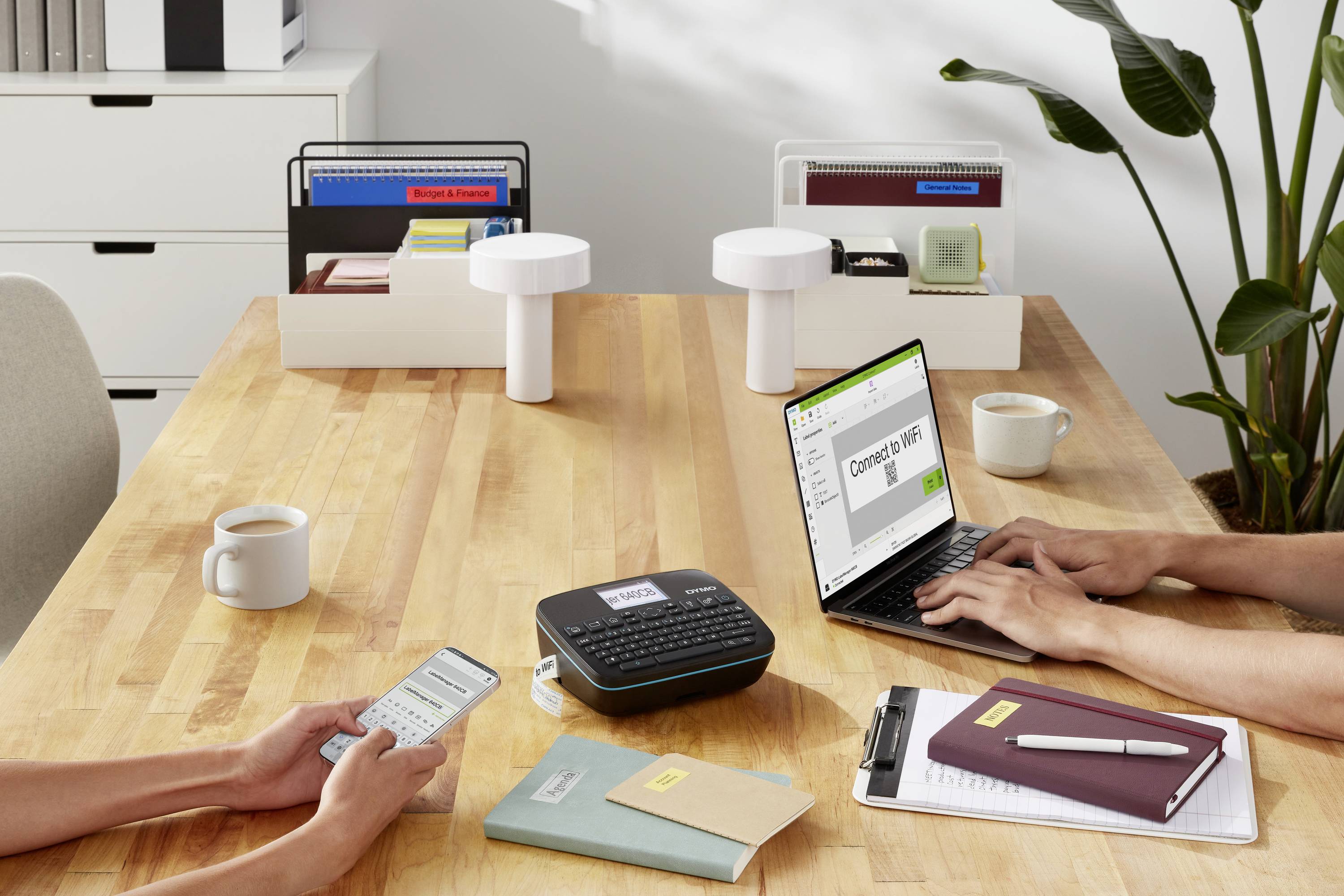 Zwei Personen sitzen an einem Holztisch. Eine Person benutzt einen Laptop, die andere ein Smartphone. Auf dem Tisch stehen Büroordner, eine Tasse und Pflanzen.