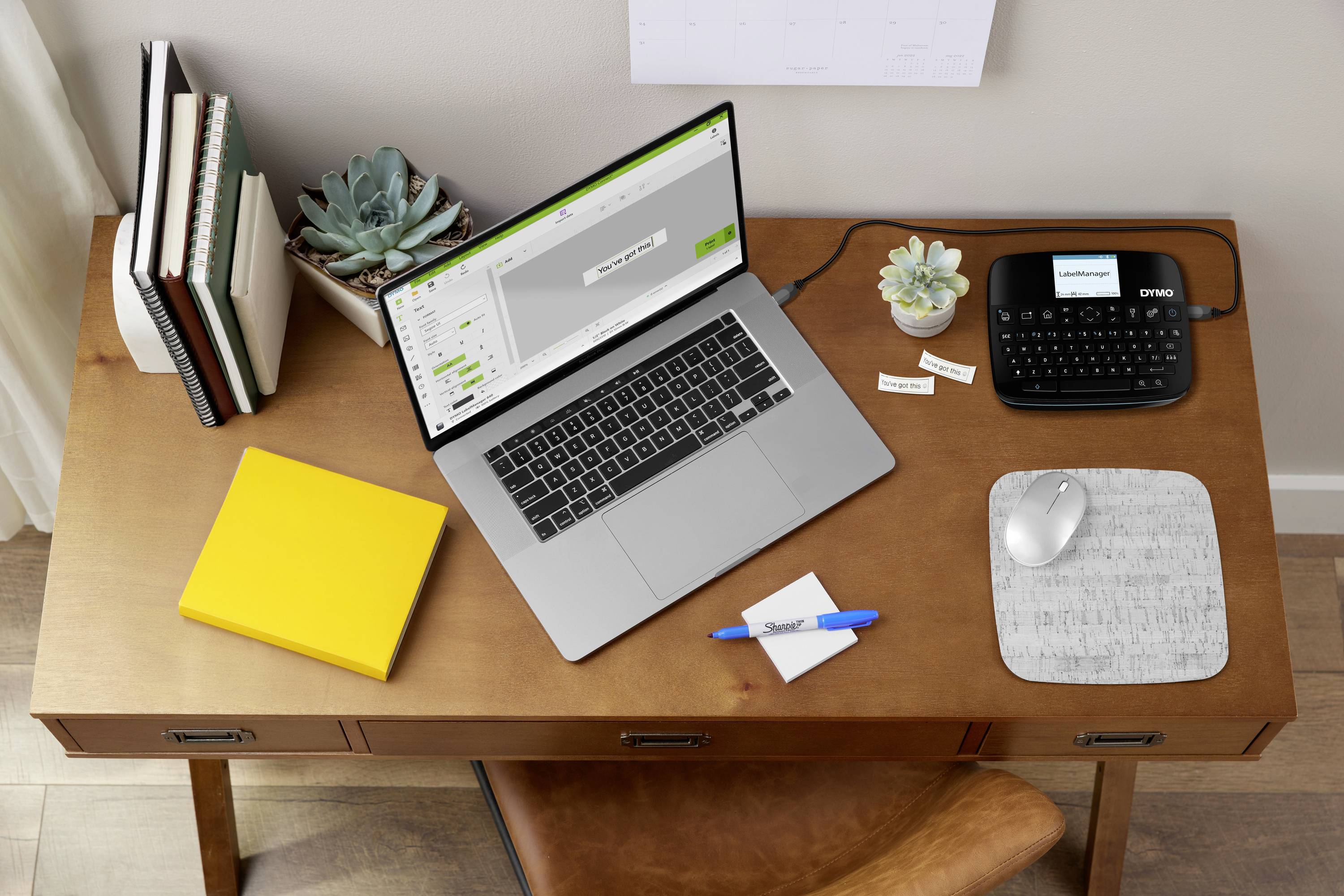 Laptop und Etikettendrucker auf einem Schreibtisch mit Pflanzen, Notizblock und Stift, zeigen eine organisierte Arbeitsumgebung.