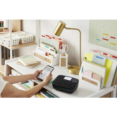 Eine Person bedient ein Etikettiergerät auf einem Schreibtisch, umgeben von organisiertem Büromaterial, einer Lampe und einem Kalender.