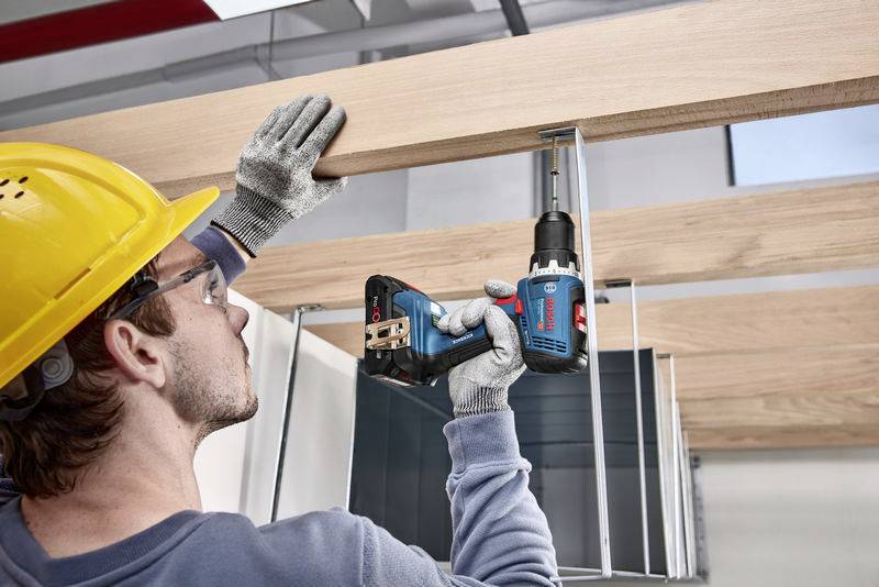 Ein Arbeiter mit Schutzbrille und Helm befestigt eine Holzplatte an einer Metallstruktur mit einem elektrischen Werkzeug.