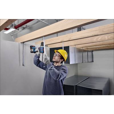 Ein Handwerker mit Helm und Schutzbrille befestigt Metallhalterungen mit einem Bohrschrauber an einer Holzdecke in einem Rohbau.