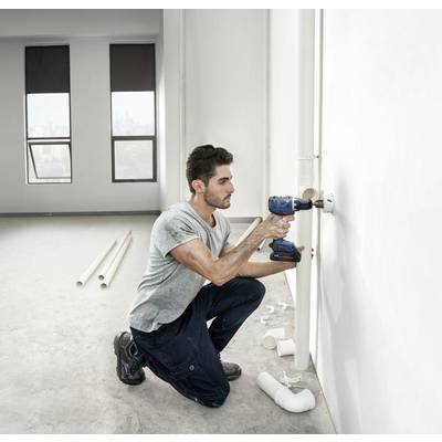 Ein Mann kniet auf dem Boden und bohrt mit einer Bohrmaschine Löcher in eine weiße Wand. Im Raum liegen Rohre und verschiedene Werkzeuge.