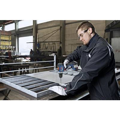 Mann in Arbeitskleidung bohrt Metall in einer Werkstatt. Er trägt Schutzbrille und Ohrstöpsel für Sicherheit.
