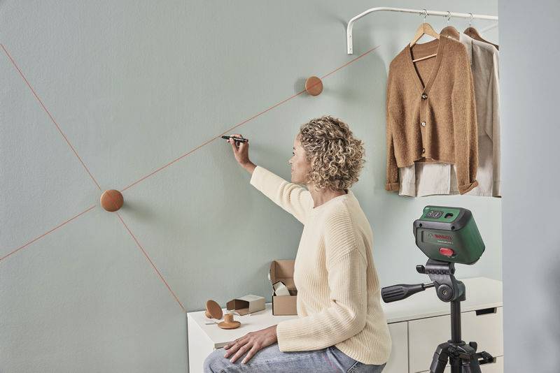 Eine Person zeichnet präzise Linien an einer Wand mit einem Laser-Nivelliergerät. Kleidung hängt im Hintergrund.