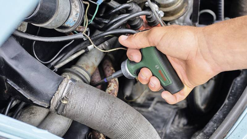 Eine Hand hält einen elektrischen Schraubenzieher und arbeitet an einem Motor mit mehreren Kabeln und Schläuchen.