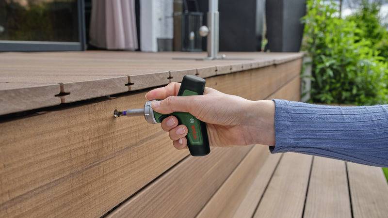 Eine Hand hält einen elektrischen Schraubendreher und zieht eine Schraube an einer Holzterrasse fest. Im Hintergrund sind Pflanzen sichtbar.