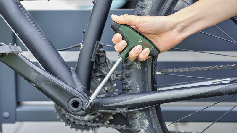Eine Hand zieht mit einem Inbusschlüssel eine Schraube am Rahmen eines schwarzen Fahrrads fest. Das Fahrrad ist auf einem Ständer.