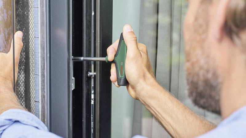 Eine Person repariert ein Fensterrahmen mit einem Schraubendreher, hält ein Gerät zur Unterstützung des Gewichts in der anderen Hand.