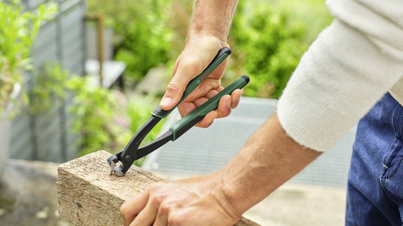 Eine Person zieht mit einer Kneifzange einen Nagel aus einem Holzstück. Die Szene befindet sich im Freien.