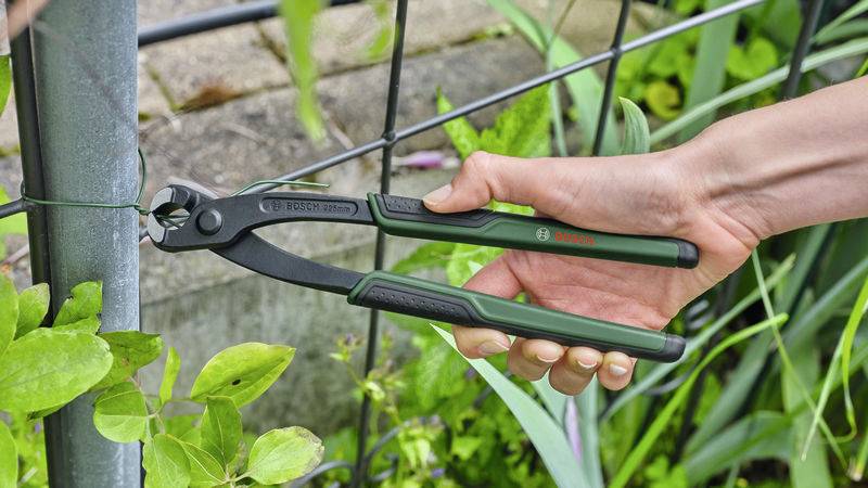 Eine Hand hält eine grüne Zange und schneidet einen Draht an einem Metallzaun. Im Hintergrund sind Pflanzen sichtbar.