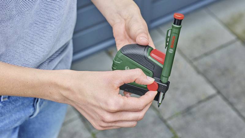 Eine Person hält ein grünes und schwarzes Werkzeug, vermutlich ein Messband mit integriertem Stift. Die Hände sind gut sichtbar.
