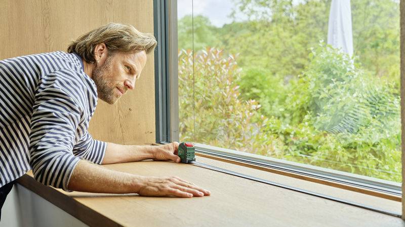 Ein Mann misst die Breite eines Fensters mit einem Laser-Entfernungsmesser. Draußen ist grünes Laub sichtbar.