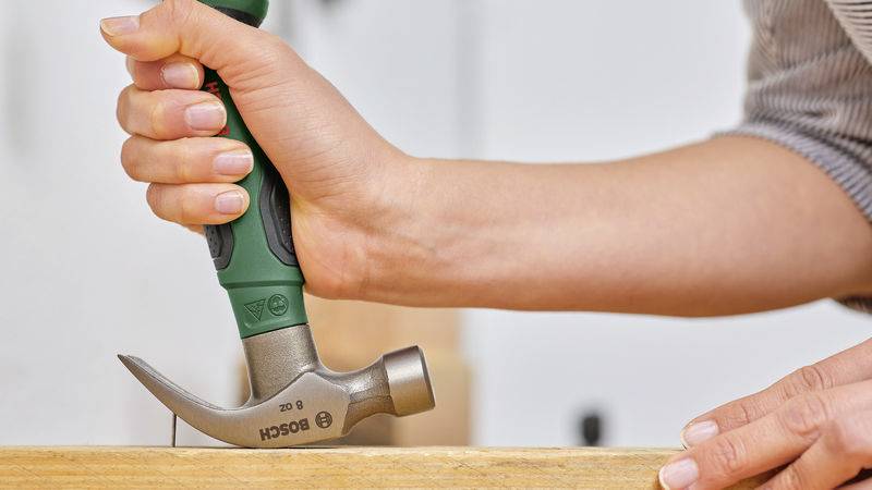 Eine Hand hält einen Hammer und schlägt einen Nagel in ein Holzstück. Die Szene zeigt die Ausführung handwerklicher Arbeit.