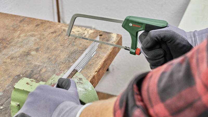 Eine Person sägt mit einer Handsäge ein Metallstück, das in einem Schraubstock befestigt ist. Sie trägt Arbeitshandschuhe.